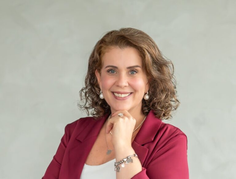 Natália Sansoni, superintendente de Marketing, Comunicação e Experiência do Cliente, é loira, cabelos no ombro, está de camiseta branca e blaser vermelho